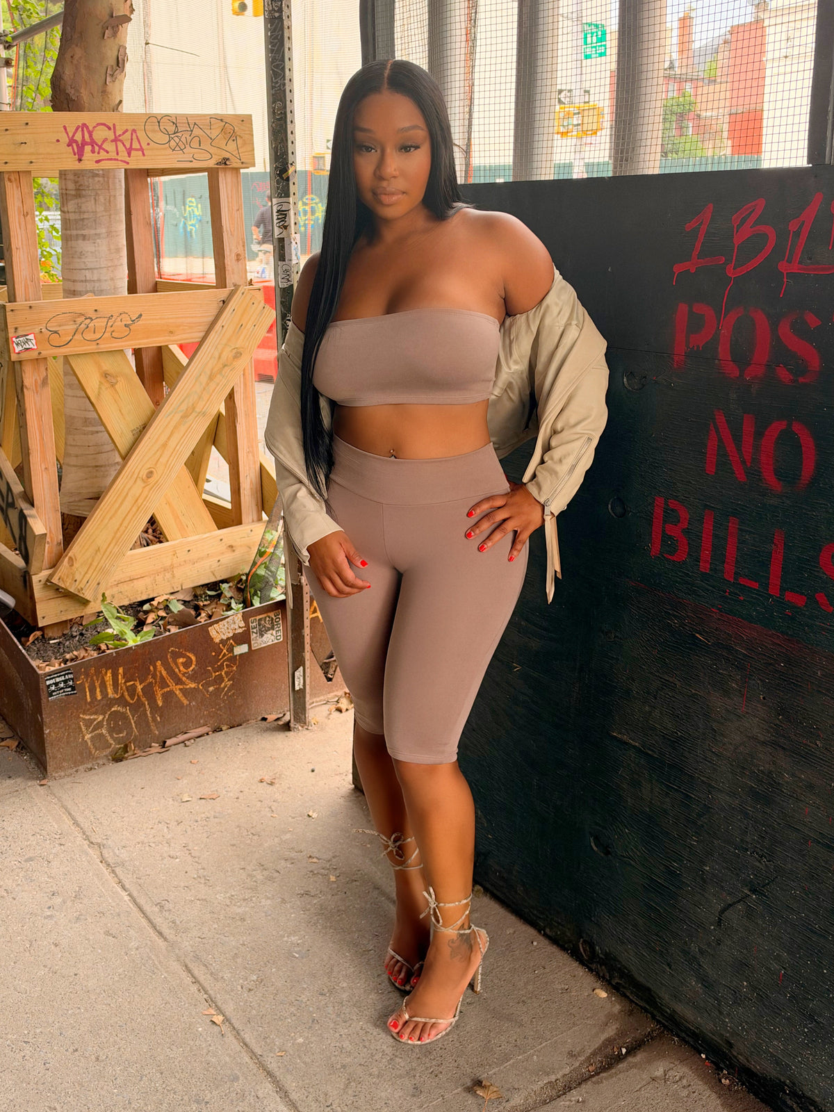 A woman posing on a city sidewalk wearing a matching beige tube top and biker shorts set, with a light beige cardigan draped off her shoulders and gold high-heeled sandals.