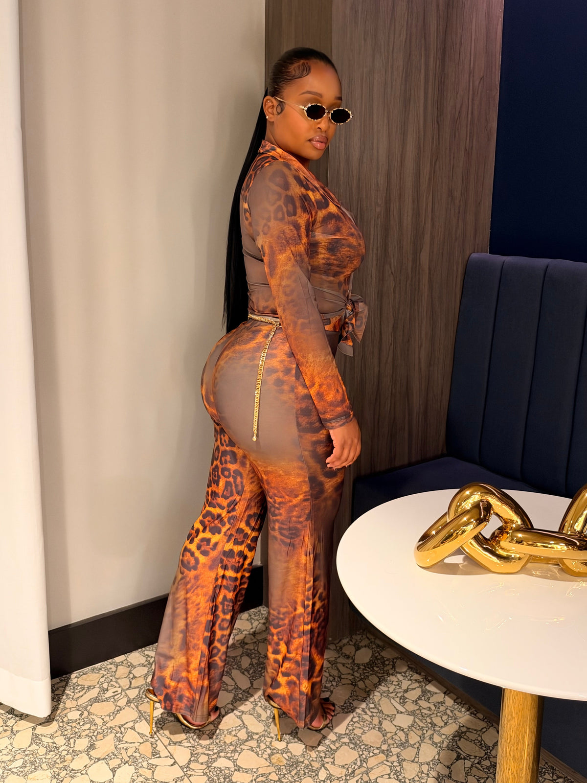 A woman posing in a matching sheer orange and black leopard-print jumpsuit with a gold zipper detail, standing in a modern indoor setting with a white curtain, wood paneling, and a blue wall.