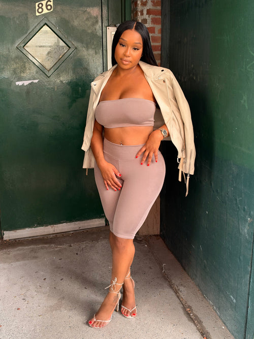 A woman models a matching beige two-piece activewear set consisting of a bandeau crop top and high-waisted shorts, styled with an open cream jacket and strappy heels, standing in front of a green door and brick wall.
