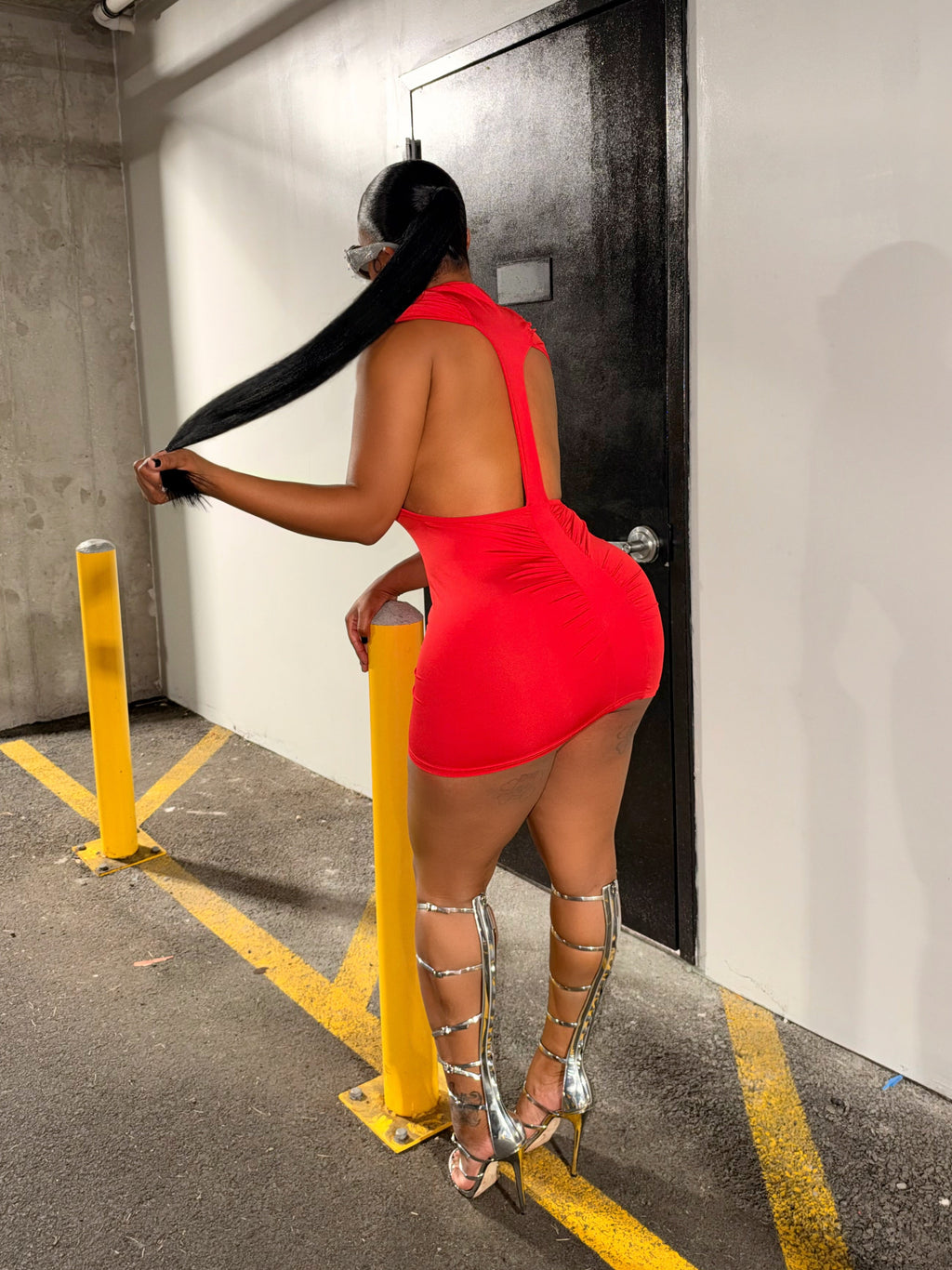 A woman stands in a parking garage wearing a bright red backless mini dress and silver strappy high-heeled sandals, posing near yellow bollards and a black door.