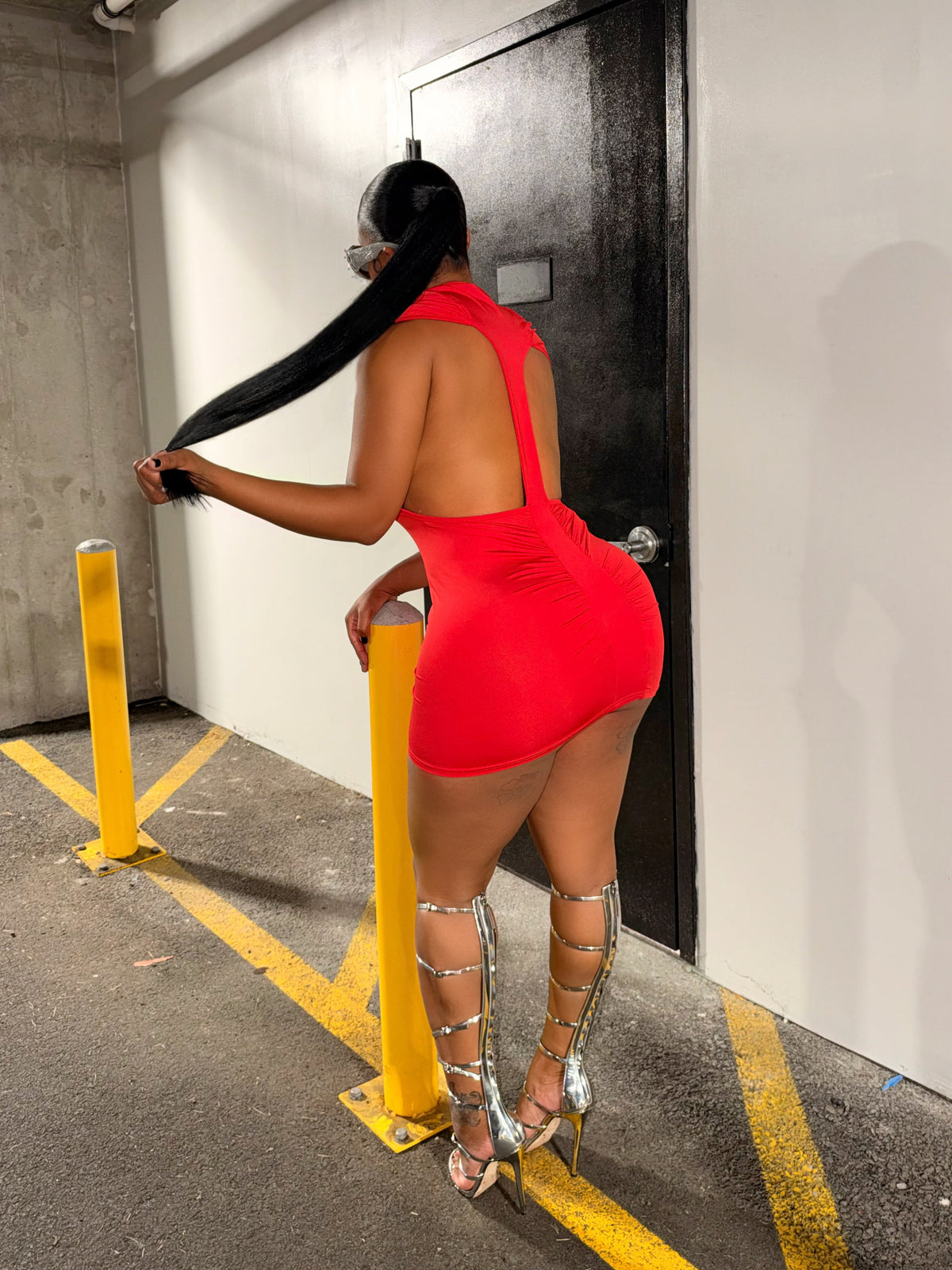 A woman stands in a parking garage wearing a bright red backless mini dress and silver strappy high-heeled sandals, posing near yellow bollards and a black door.