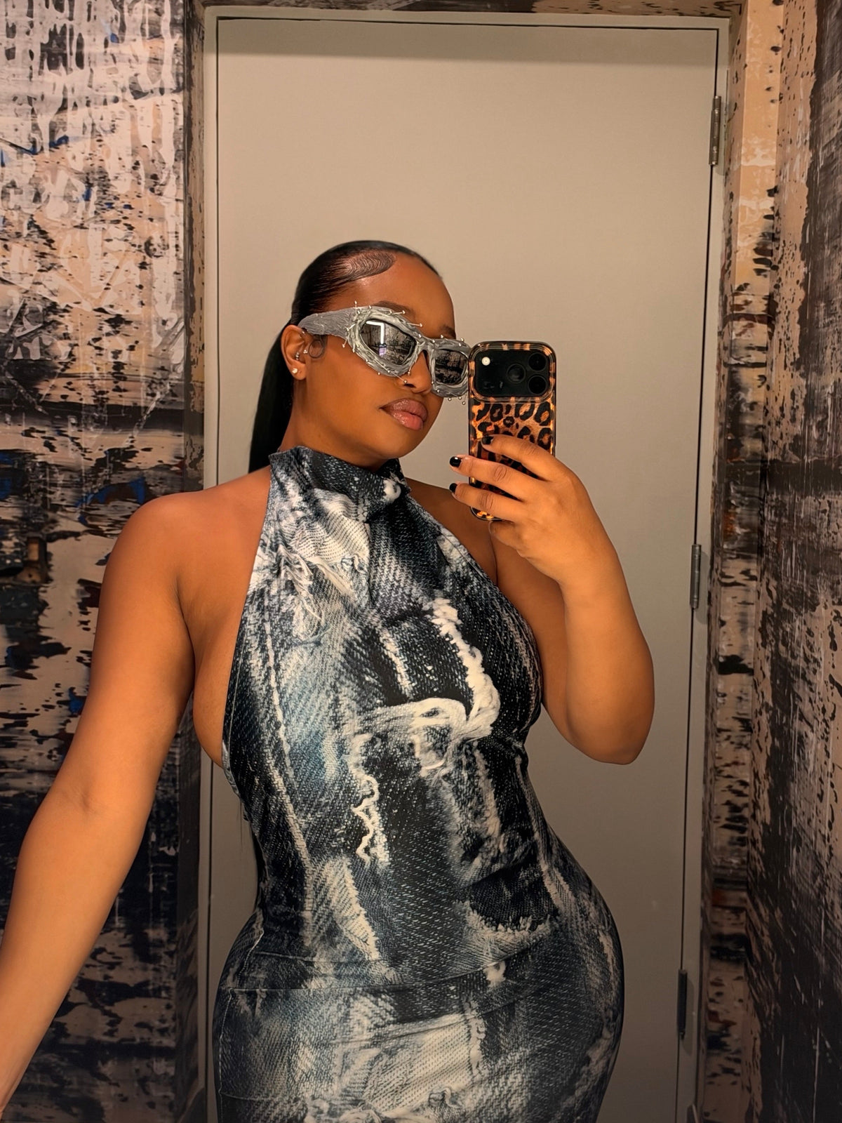 A woman takes a mirror selfie wearing a high-neck, bodycon halter dress with a black and white snakeskin pattern, along with silver-framed sunglasses.