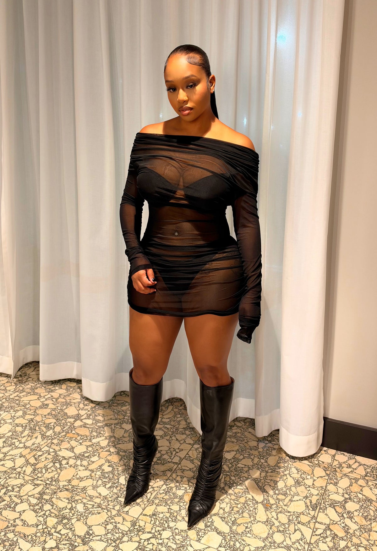 A woman stands in front of white curtains wearing a sheer black off-the-shoulder mini dress with long sleeves and black knee-high pointed boots.
