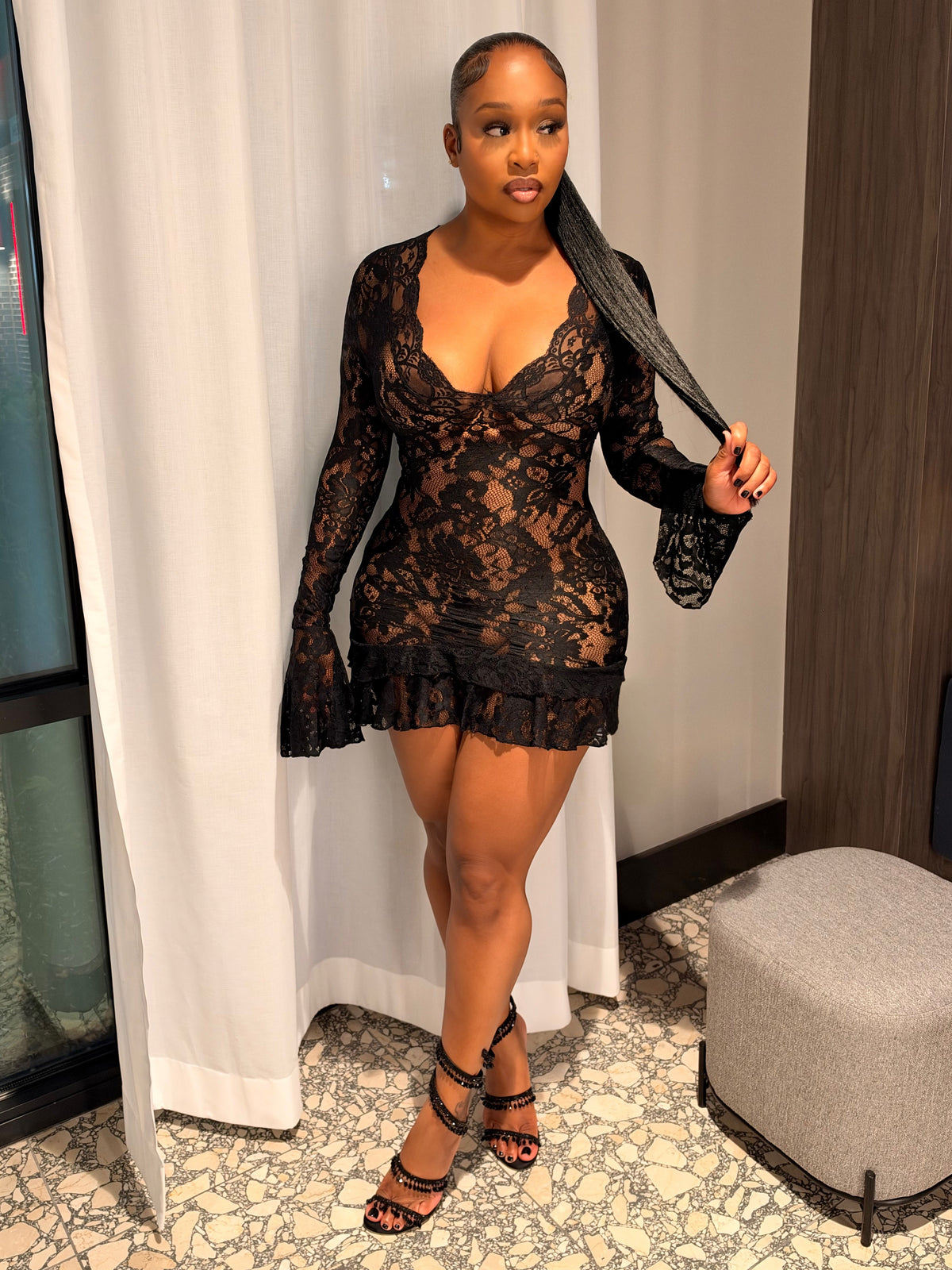 A woman models a black lace long-sleeve mini dress with a ruffled hem in an indoor setting with white curtains, a patterned floor, and a gray ottoman.