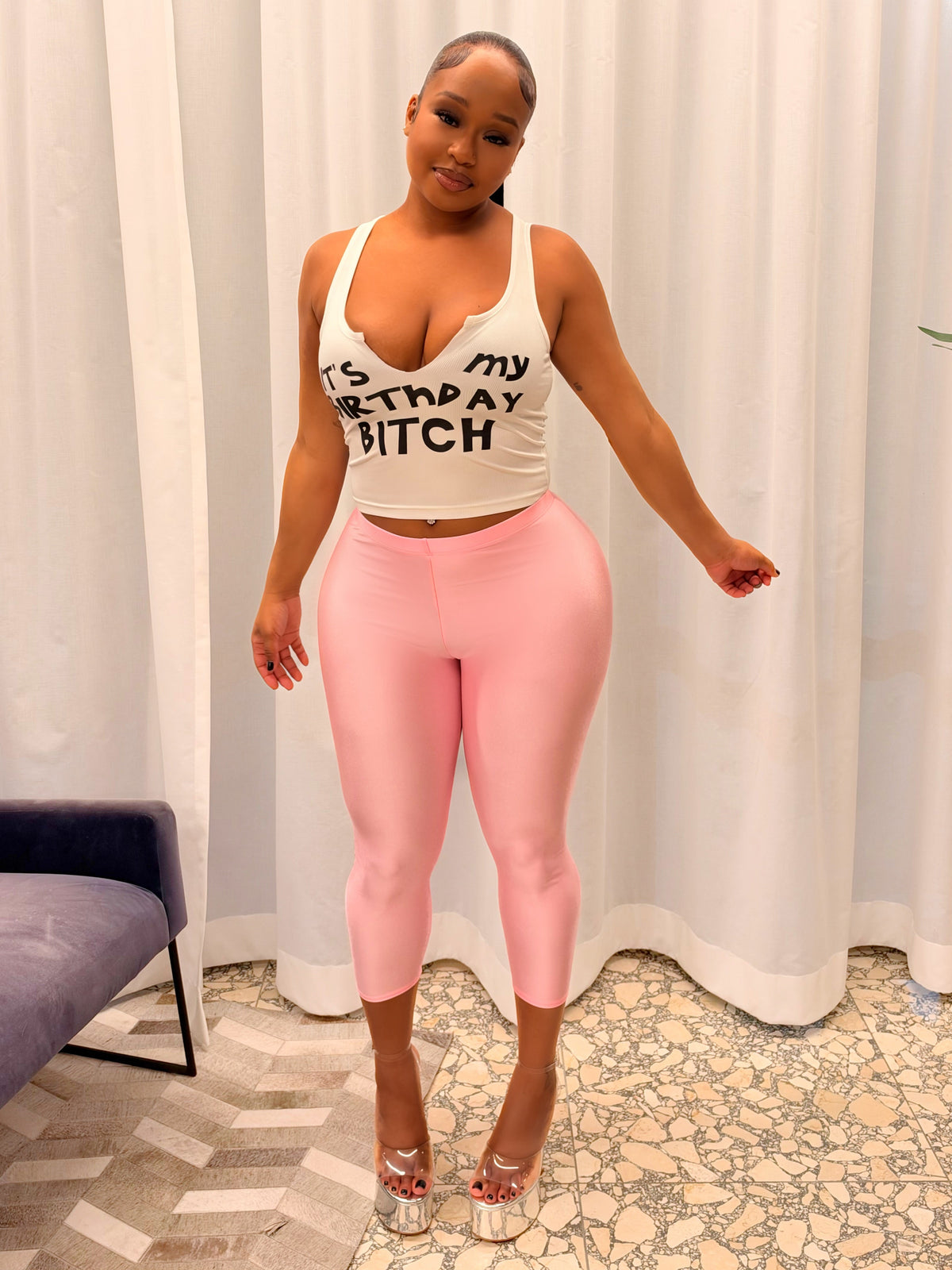 A woman modeling a two-piece outfit consisting of a white tank top with black text and pink leggings, standing in front of white curtains in an indoor setting.