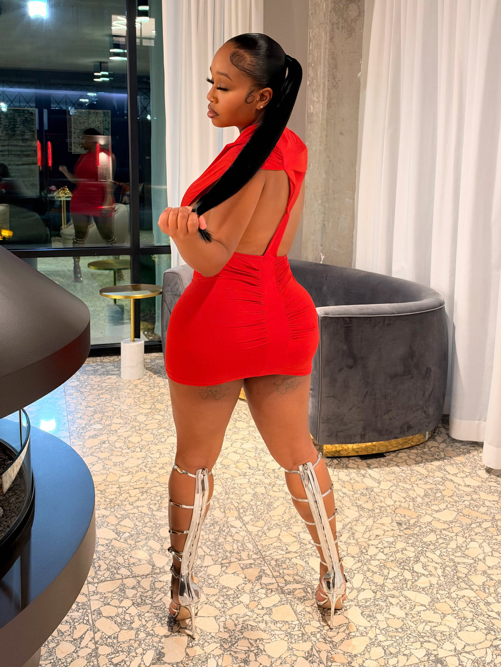 A woman stands indoors wearing a bright red mini dress with a low back, paired with metallic high-heeled boots. The setting features a modern interior with a fireplace, white curtains, and a textured floor.