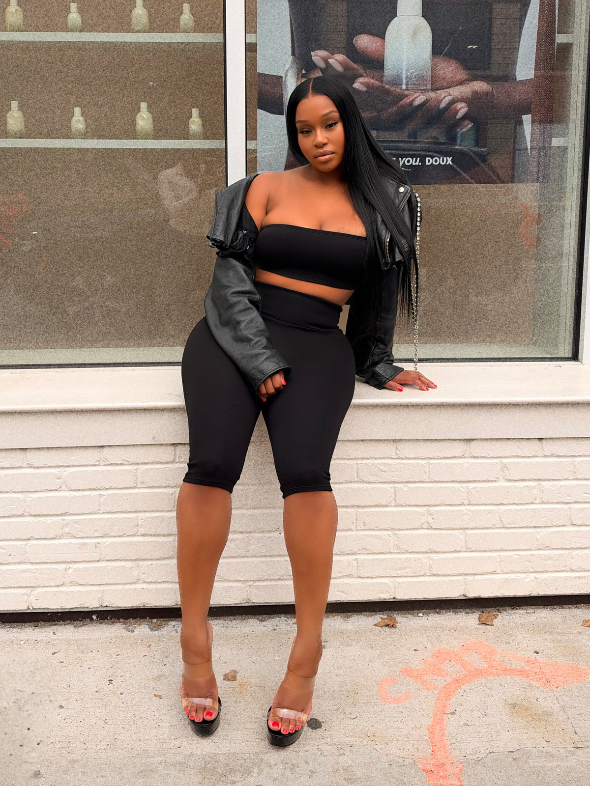 A woman posing in a black two-piece crop top and biker shorts set with a black leather jacket, standing in front of a storefront window.