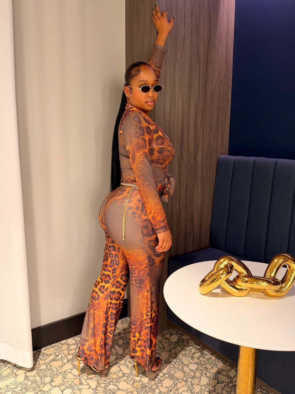 A woman posing in a matching sheer orange and brown leopard-print jumpsuit with a gold zipper detail, standing in a modern indoor setting with a blue wall, wooden paneling, and a small round table with a gold chain sculpture.
