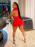 A woman posing in a bright red backless mini dress with white strappy high heels inside a modern room with a fireplace and white curtains.