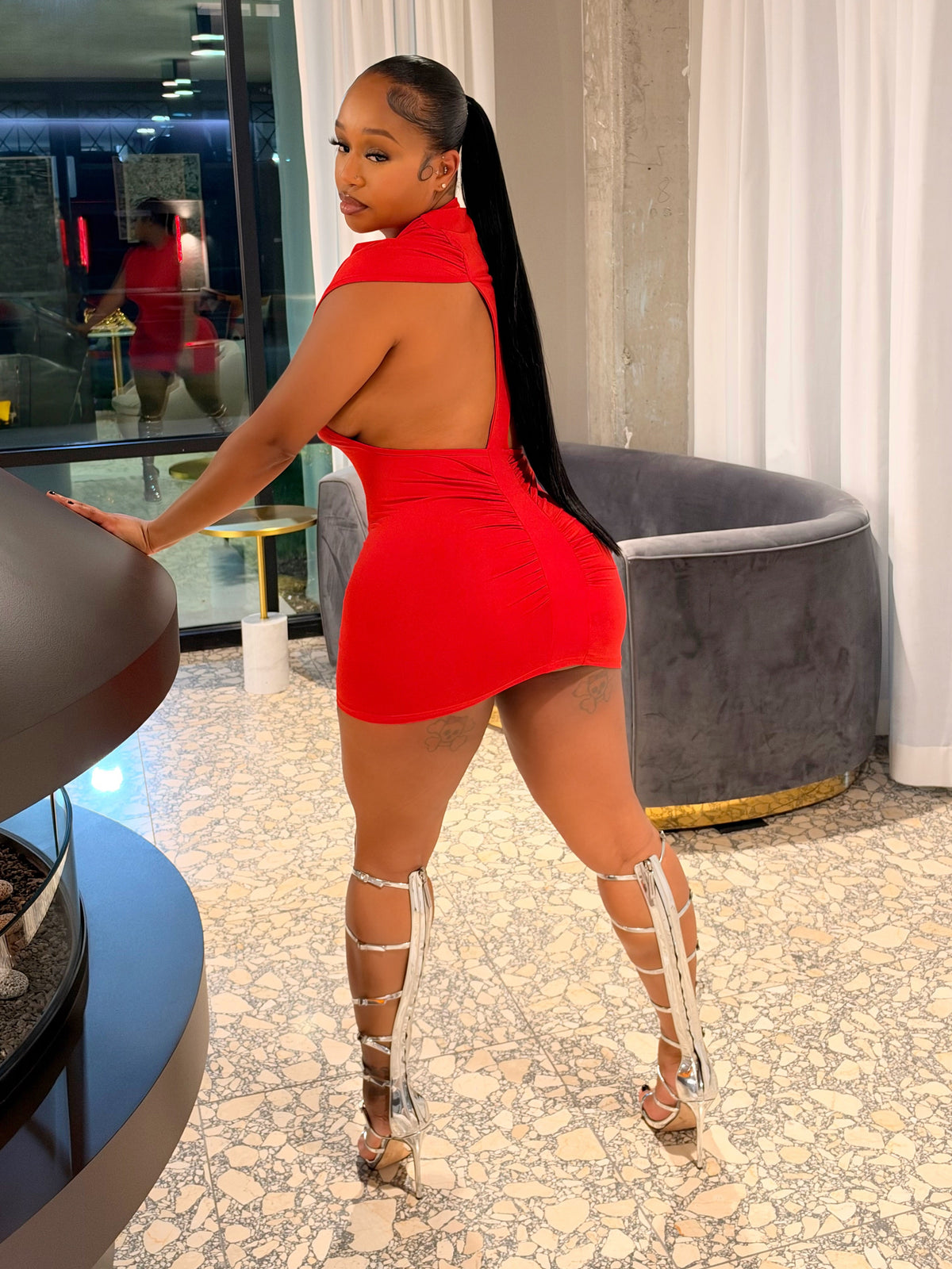 A woman posing in a bright red backless mini dress with white strappy high heels inside a modern room with a fireplace and white curtains.