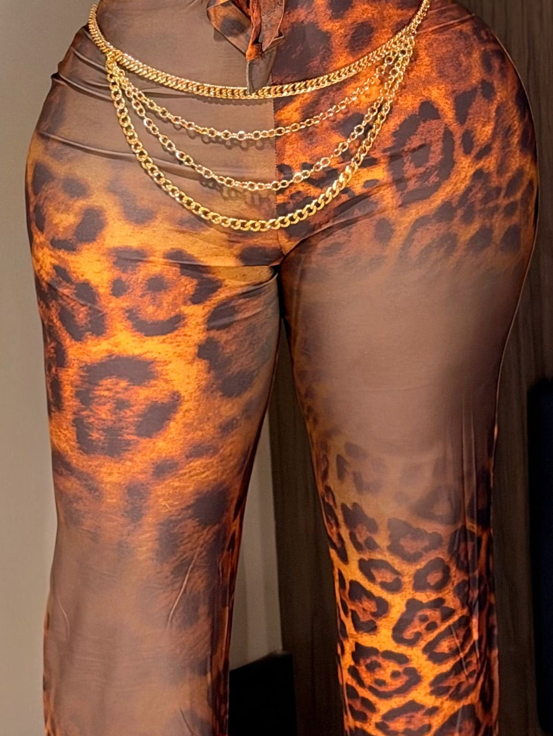 A woman modeling a sheer, long-sleeve jumpsuit with a brown, orange, and black leopard print pattern, standing in an indoor setting with a dark blue wall and a white table visible.