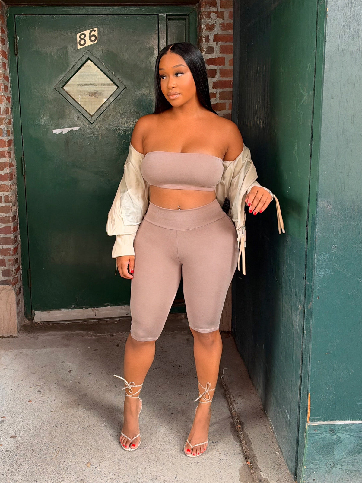 A woman stands outdoors in front of a green door and brick wall, wearing a matching beige bandeau top and biker shorts set with a cream draped cardigan and strappy high-heeled sandals.