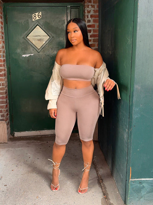 A woman stands outdoors in front of a green door and brick wall, wearing a matching beige bandeau top and biker shorts set with a cream draped cardigan and strappy high-heeled sandals.