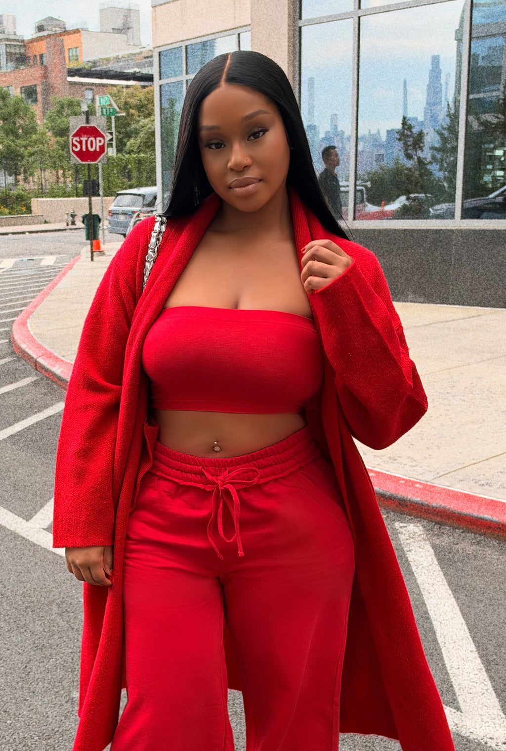 A woman stands on an outdoor city street wearing a bright red matching outfit consisting of a strapless tube top, wide-leg drawstring pants, and a long open-front coat. The background shows a modern glass building, parked cars, and a paved road.