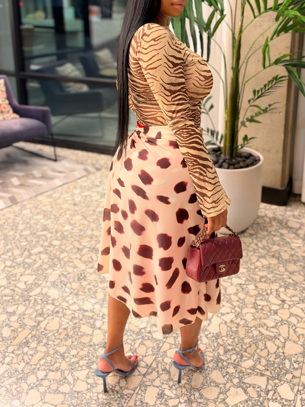 A woman posing in a modern indoor setting, wearing a beige and brown zebra-print long-sleeve top paired with a pink and brown spotted midi skirt, holding a burgundy handbag.