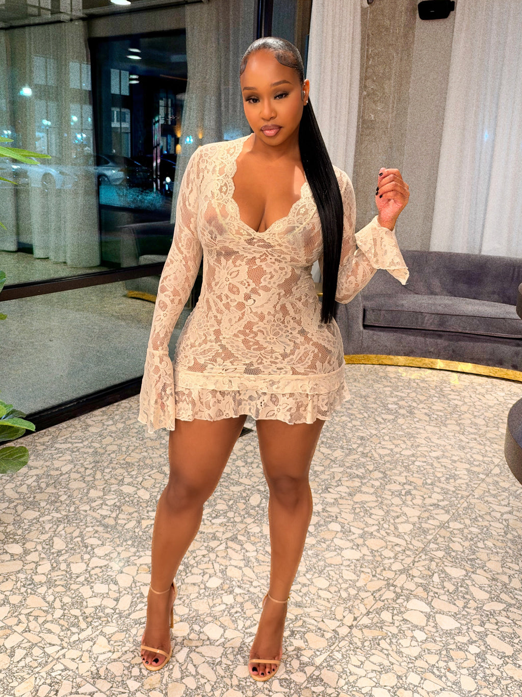 A woman posing in a beige lace mini dress with long sleeves and ruffled trim, standing in a modern indoor setting with a patterned floor, glass windows, and a plant.