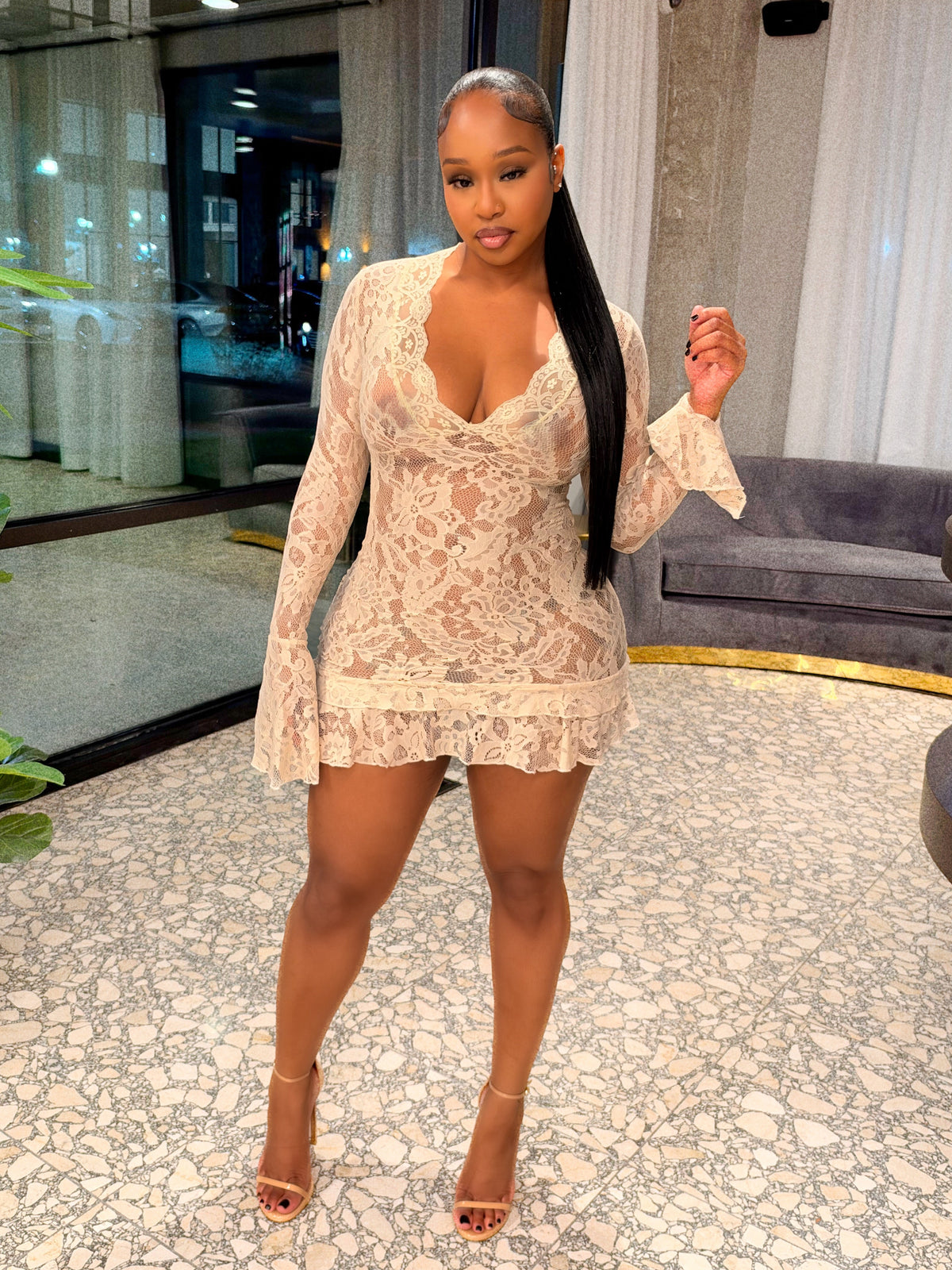 A woman posing in a beige lace mini dress with long sleeves and ruffled trim, standing in a modern indoor setting with a patterned floor, glass windows, and a plant.
