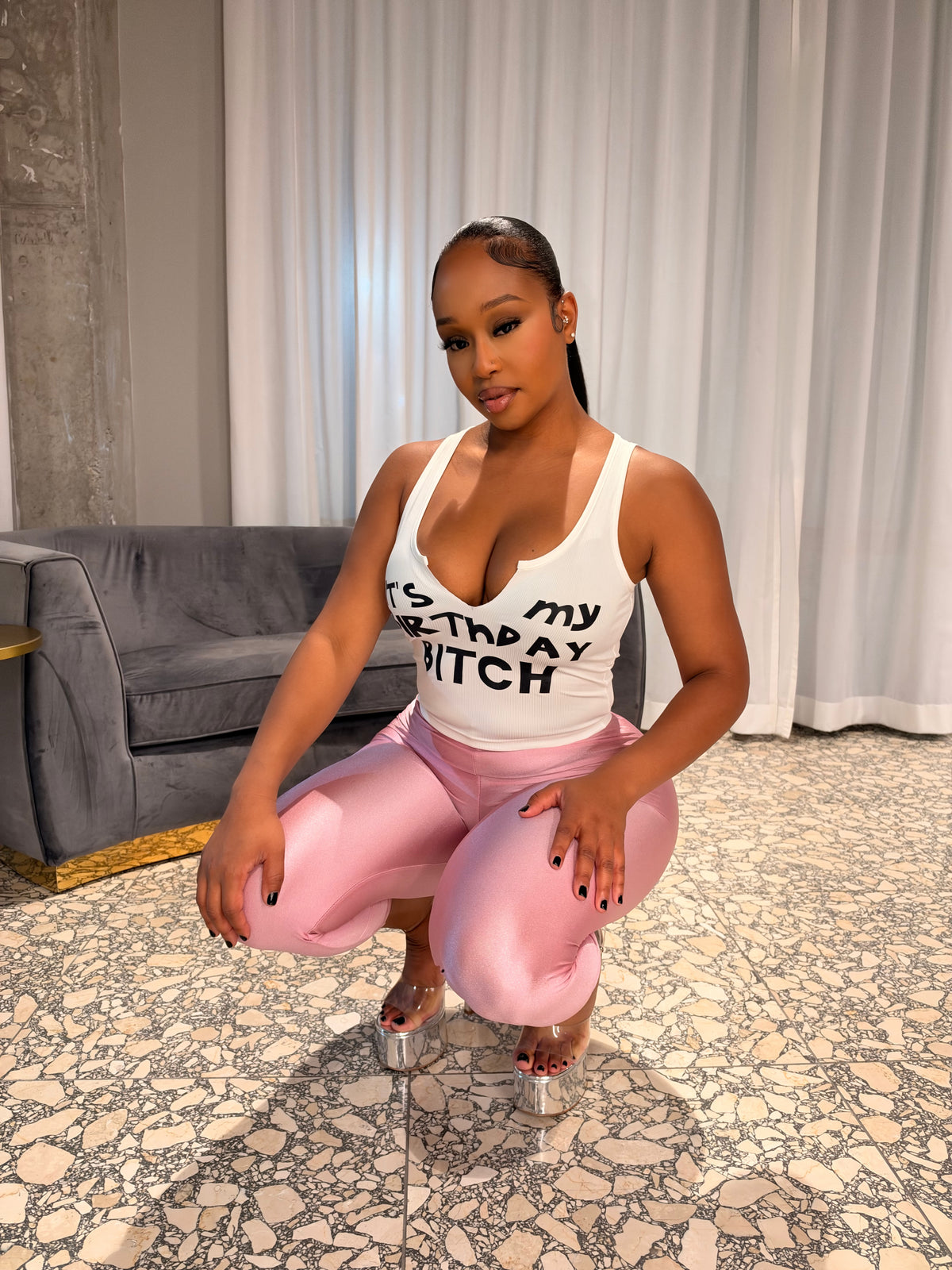 A woman crouching indoors wearing a white tank top with black text that reads 'IT'S MY BIRTHDAY BITCH' and shiny pink leggings. She is posed in front of white curtains and a gray sofa on a patterned tile floor.