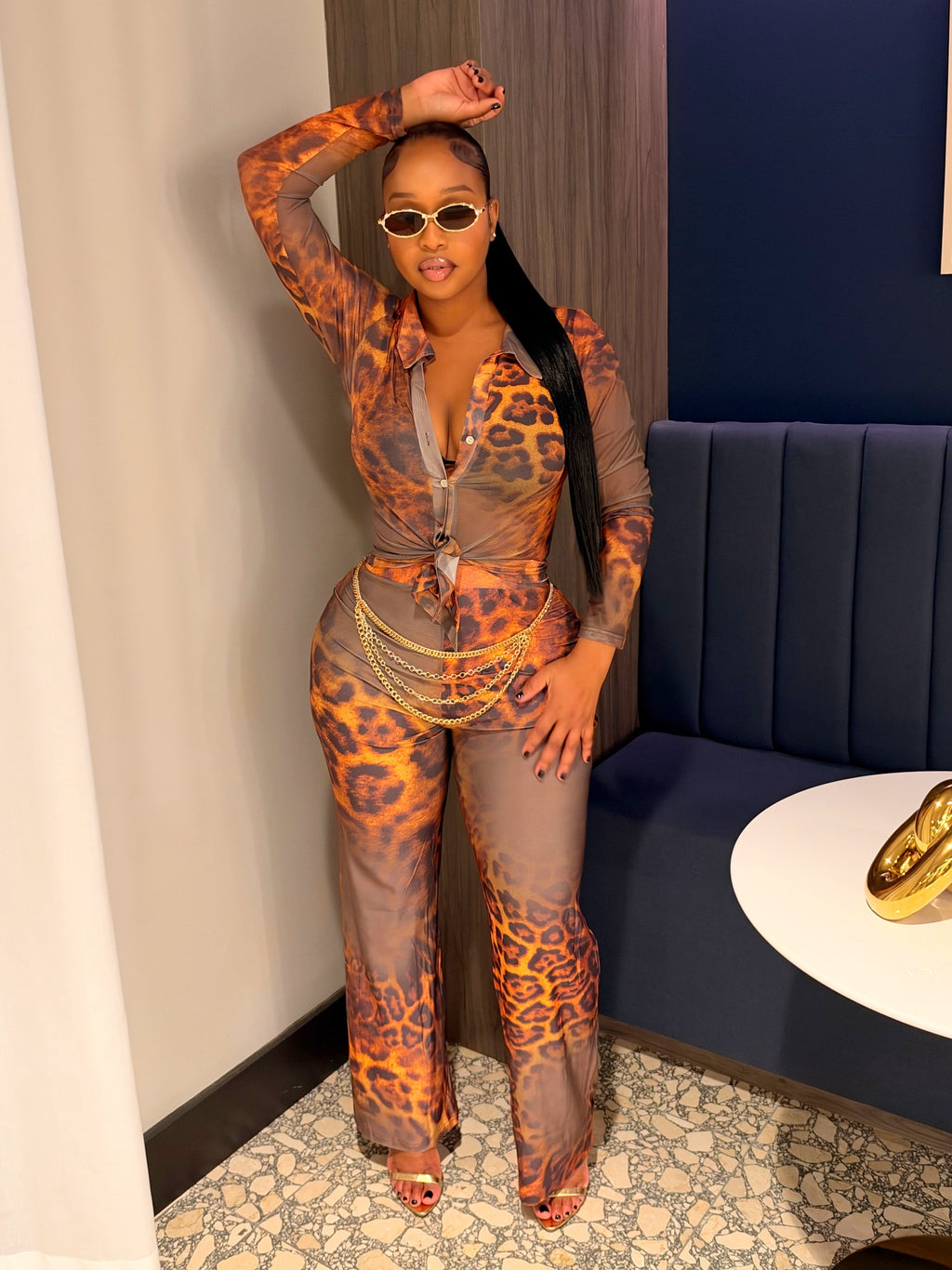 A woman posing in a matching sheer leopard-print jumpsuit with orange and brown spots over a gray base, styled with sunglasses and gold necklaces in an indoor setting.