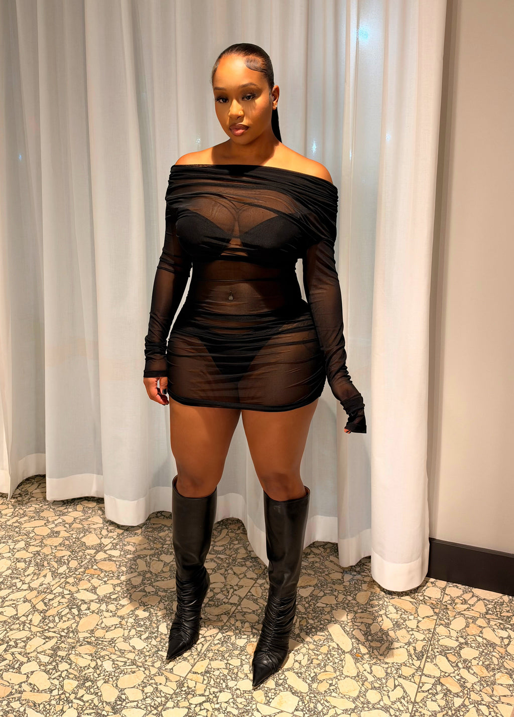 A woman stands indoors wearing a sheer black off-the-shoulder mini dress with long sleeves and matching black gloves, paired with black knee-high boots. She is posed in front of white curtains and a patterned floor.