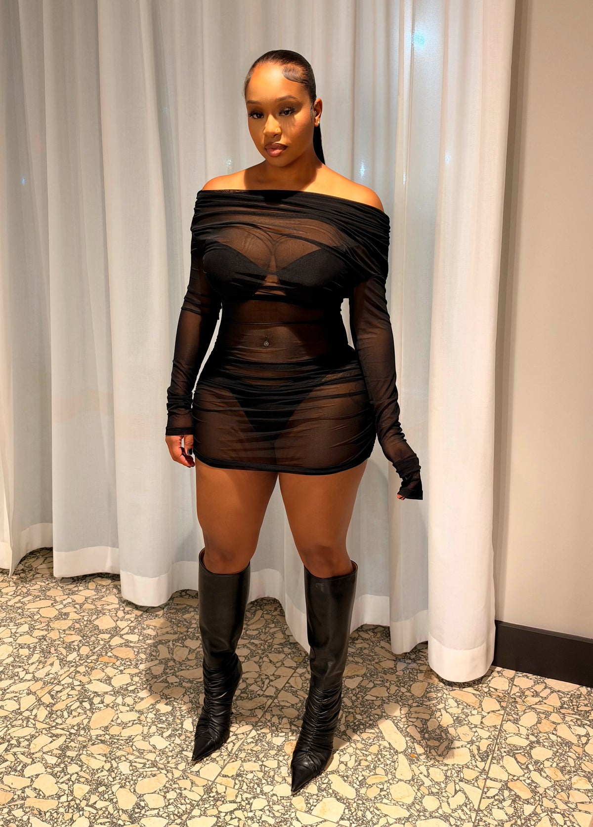 A woman stands indoors wearing a sheer black off-the-shoulder mini dress with long sleeves and matching black gloves, paired with black knee-high boots. She is posed in front of white curtains and a patterned floor.