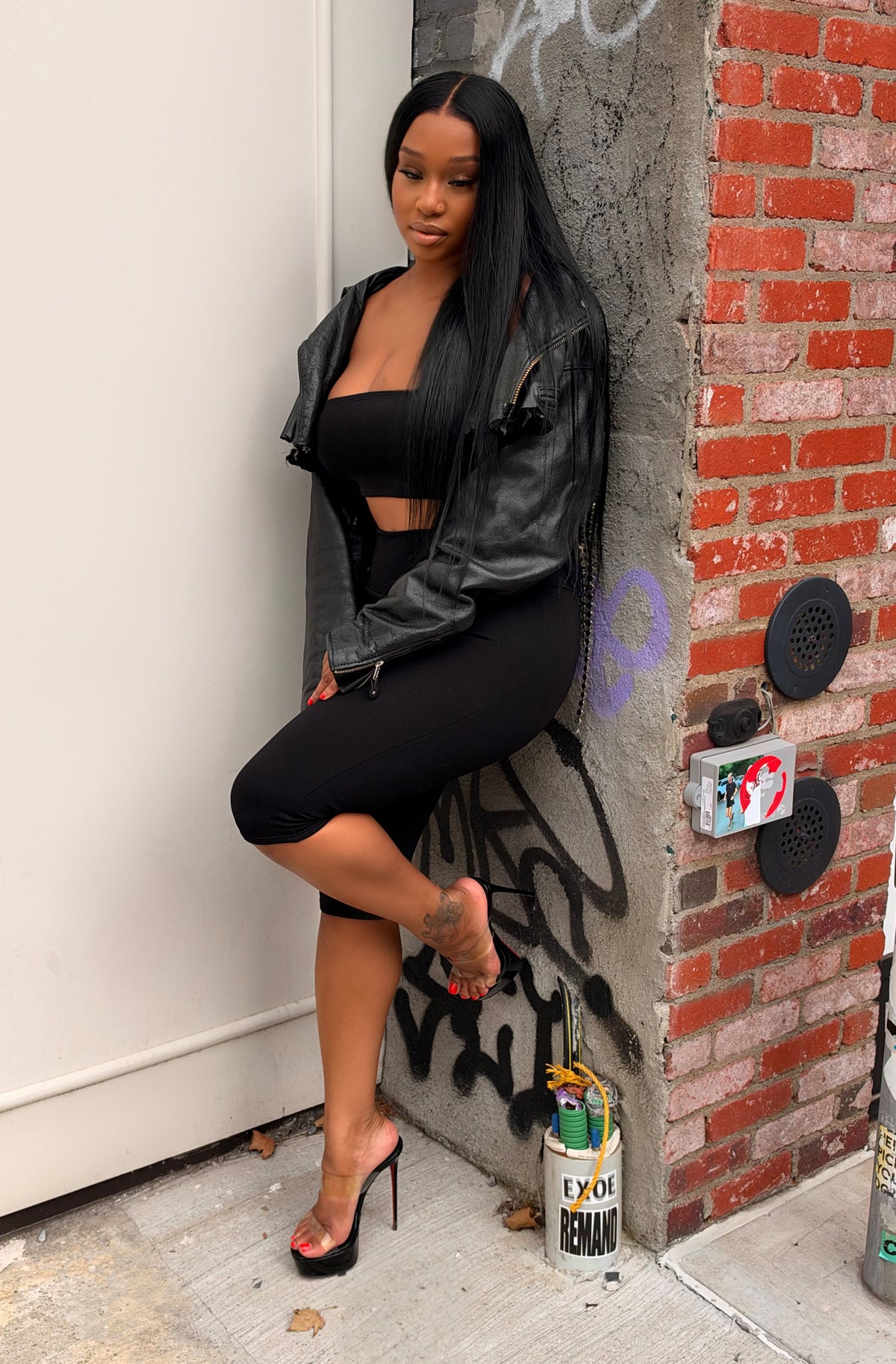 A woman posing in a black leather jacket, black crop top, and black high-waisted mini skirt with black high heels, leaning against a brick and concrete pillar in an urban setting.