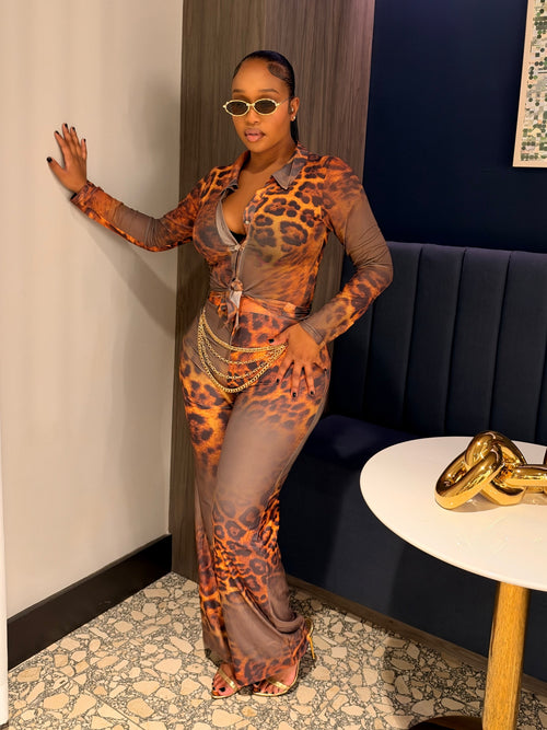 A woman posing in a matching two-piece leopard print set consisting of a sheer long-sleeve button-up shirt and matching wide-leg trousers, styled with gold necklaces and sunglasses in an indoor setting.