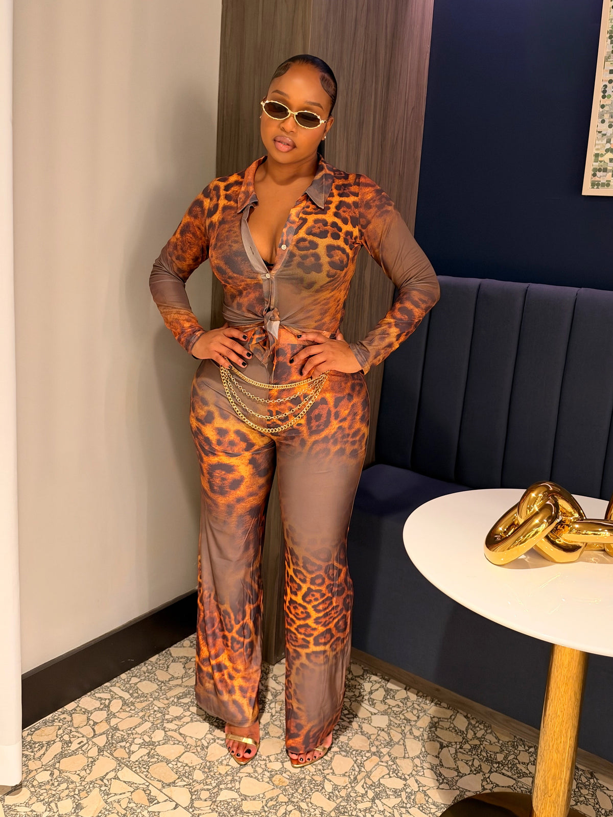 A woman wearing a sheer leopard-print jumpsuit with orange, brown, and black spots stands in an indoor setting with a dark blue wall, a white table with a gold sculpture, and a patterned floor.
