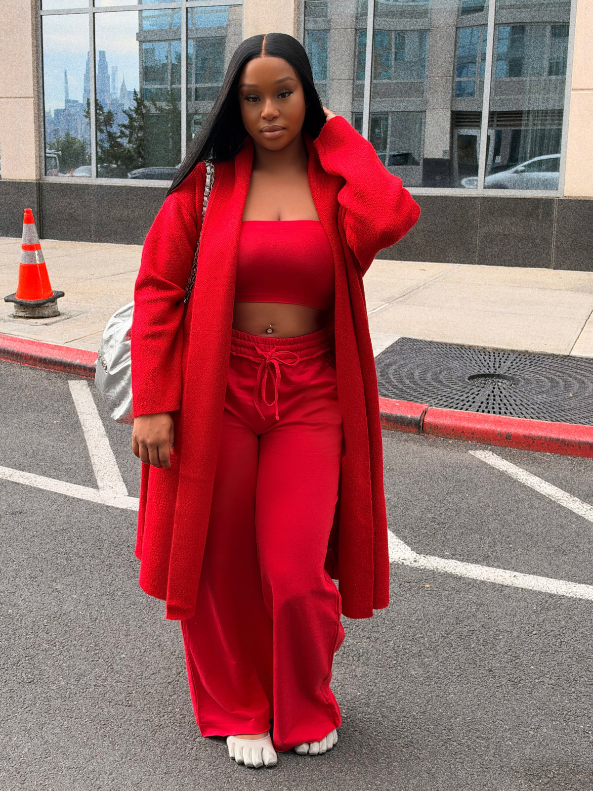 A woman stands in a parking lot wearing a bright red matching outfit consisting of a long open-front coat, cropped top, and wide-leg pants. A modern glass building is visible in the background.