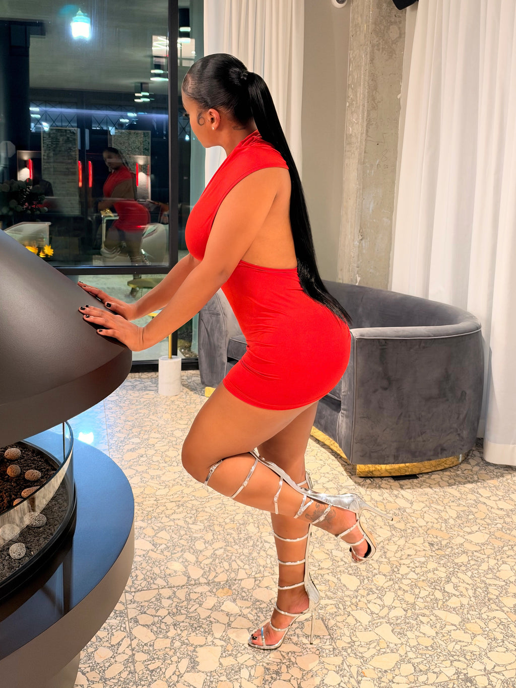 A woman in a bright red mini dress and silver strappy high heels poses next to a modern fireplace in a stylish indoor setting with white curtains and a gray lounge chair.