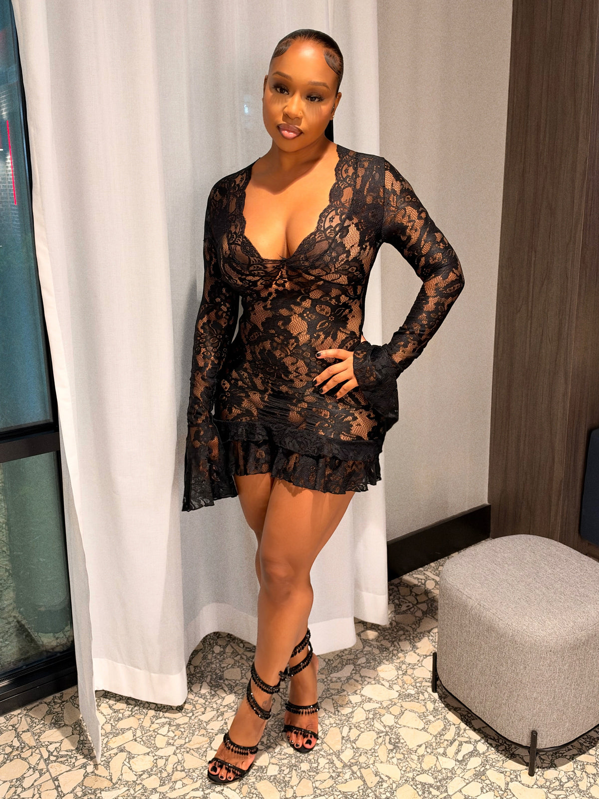 A woman posing in a black lace long-sleeve mini dress with a ruffled hem, standing in front of a white curtain in an indoor setting.
