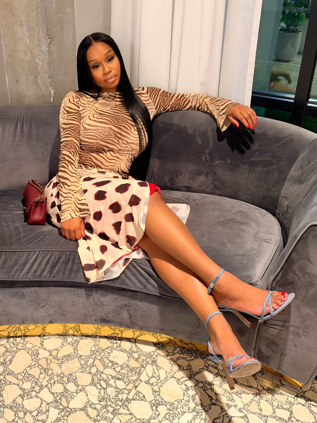 A woman posing on a gray velvet sofa wearing a beige and brown zebra-print long-sleeve top and a matching brown-and-white patterned skirt, styled with a small red handbag and blue strappy heels.