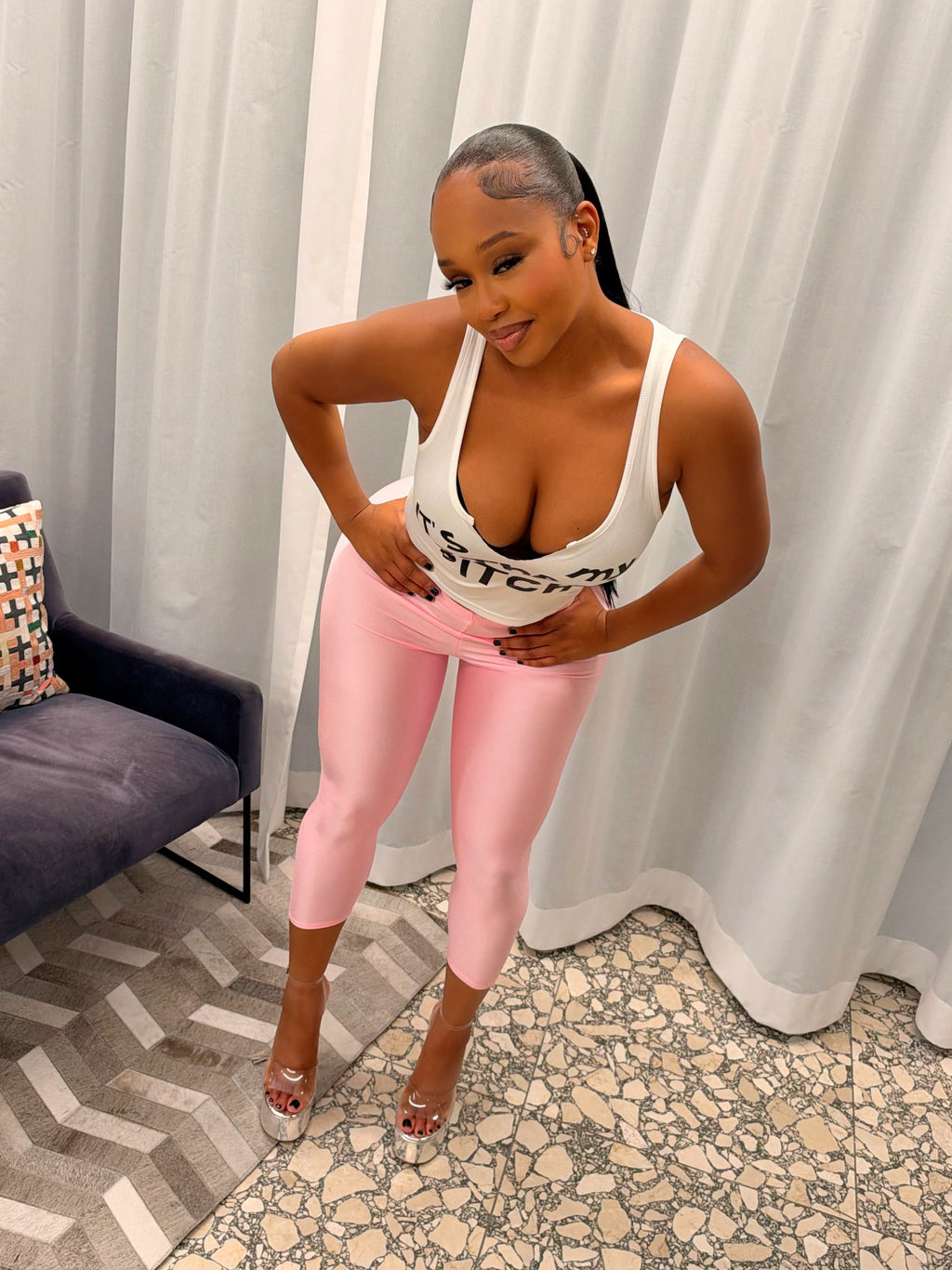 A woman posing in a white tank top with black text and light pink leggings, standing in a room with white curtains, a patterned floor, and a dark blue chair.