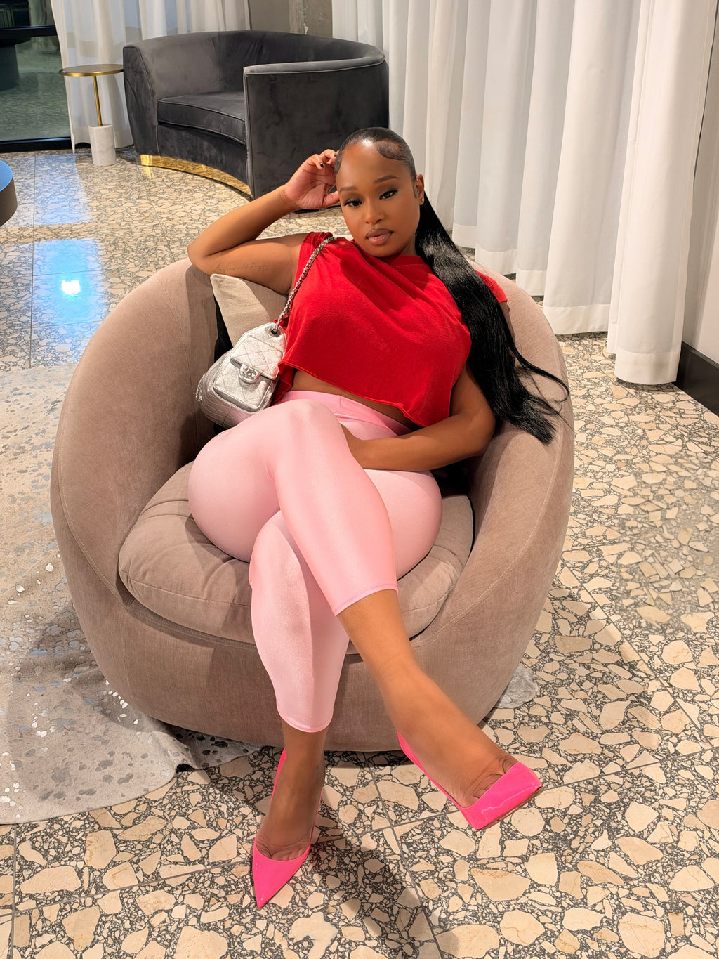 A woman posing in a modern lounge setting, wearing a vibrant red crop top, light pink pants, and hot pink pointed-toe heels while sitting in a round beige armchair.