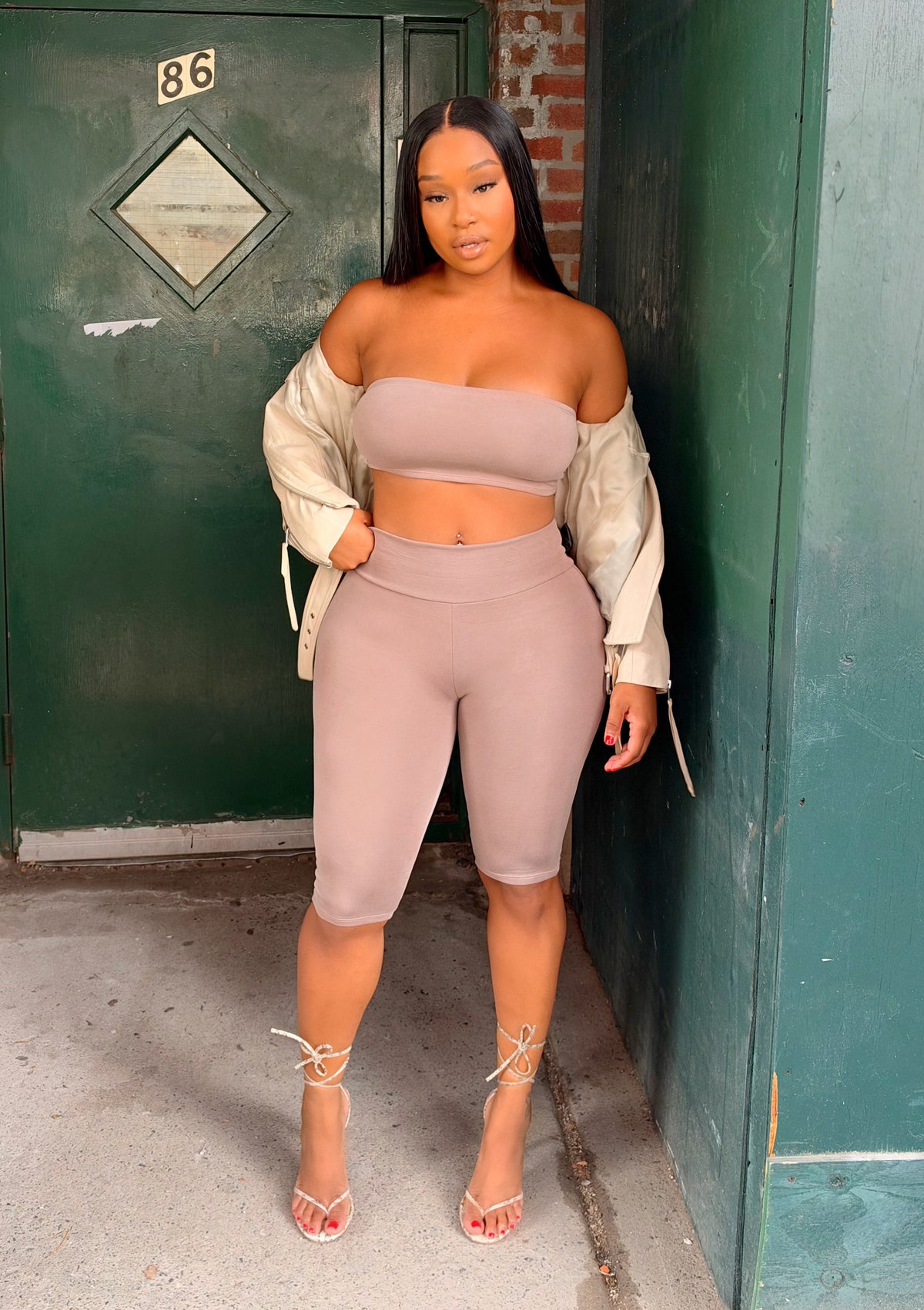 A woman stands outdoors in a narrow alleyway wearing a matching beige two-piece set consisting of an off-the-shoulder bandeau top and high-waisted biker shorts, styled with a cream jacket and strappy heels.