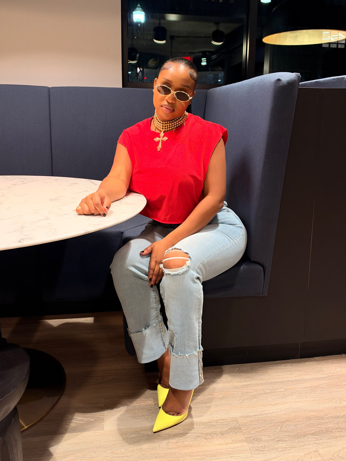 A woman sitting in a modern booth wearing a bright red sleeveless top, light-wash ripped jeans, and neon yellow pointed shoes.