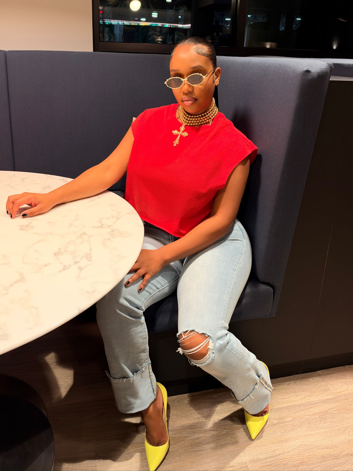 A woman sitting in a dark blue booth at a restaurant, wearing a bright red sleeveless top, light-wash ripped jeans, and neon yellow heels.