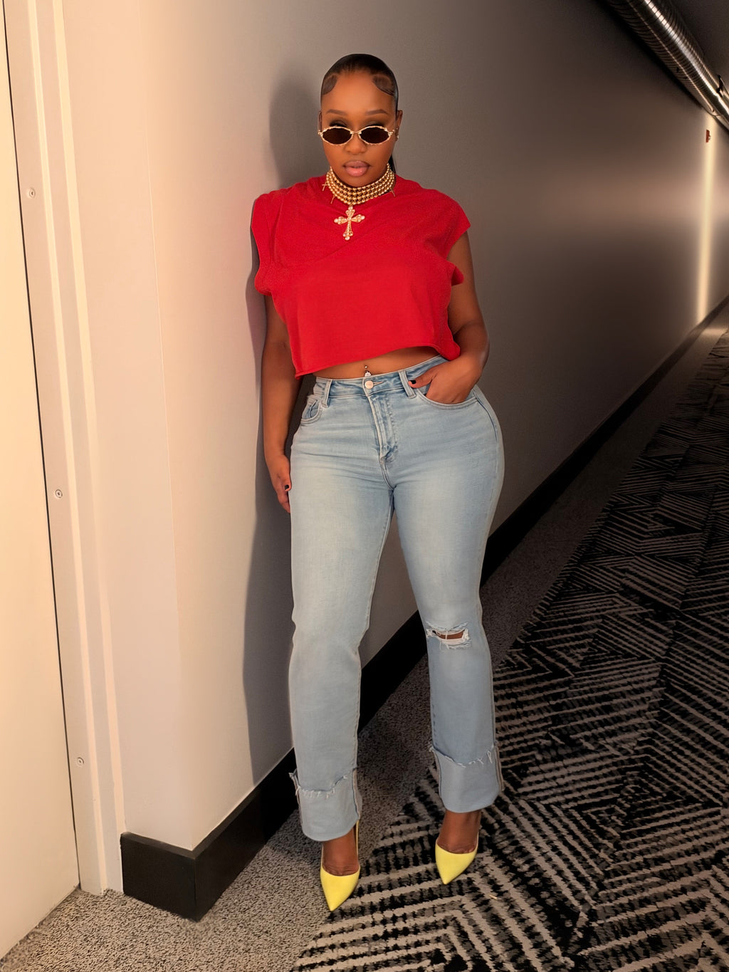 A woman posing in a hallway wearing a bright red short-sleeve crop top, light-wash distressed jeans, and yellow pointed-toe heels.