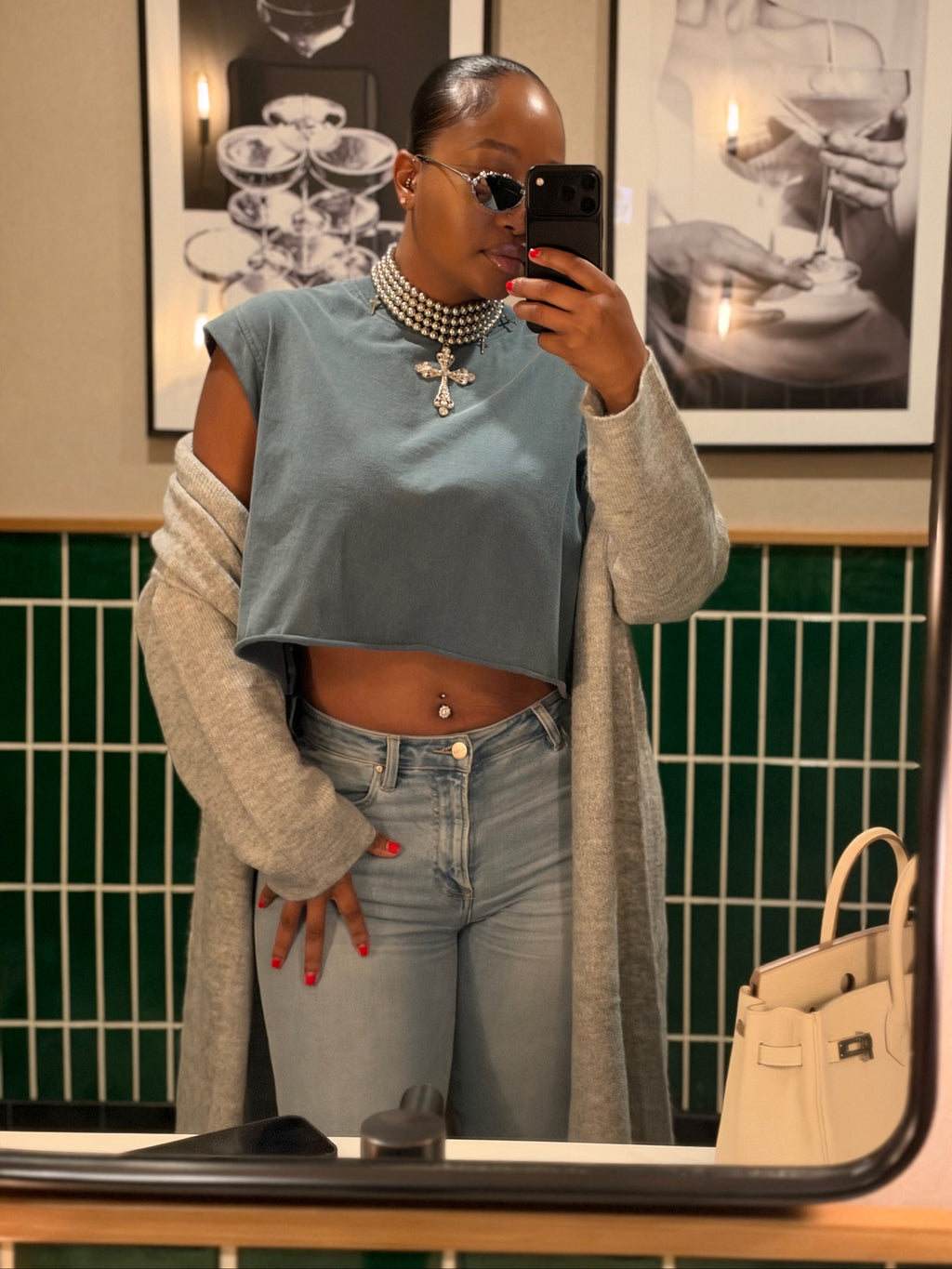 A woman takes a mirror selfie showing off a light blue cropped top with a gray cardigan, light-wash jeans, and a pearl necklace. The background features a green tiled wall and framed black-and-white art.
