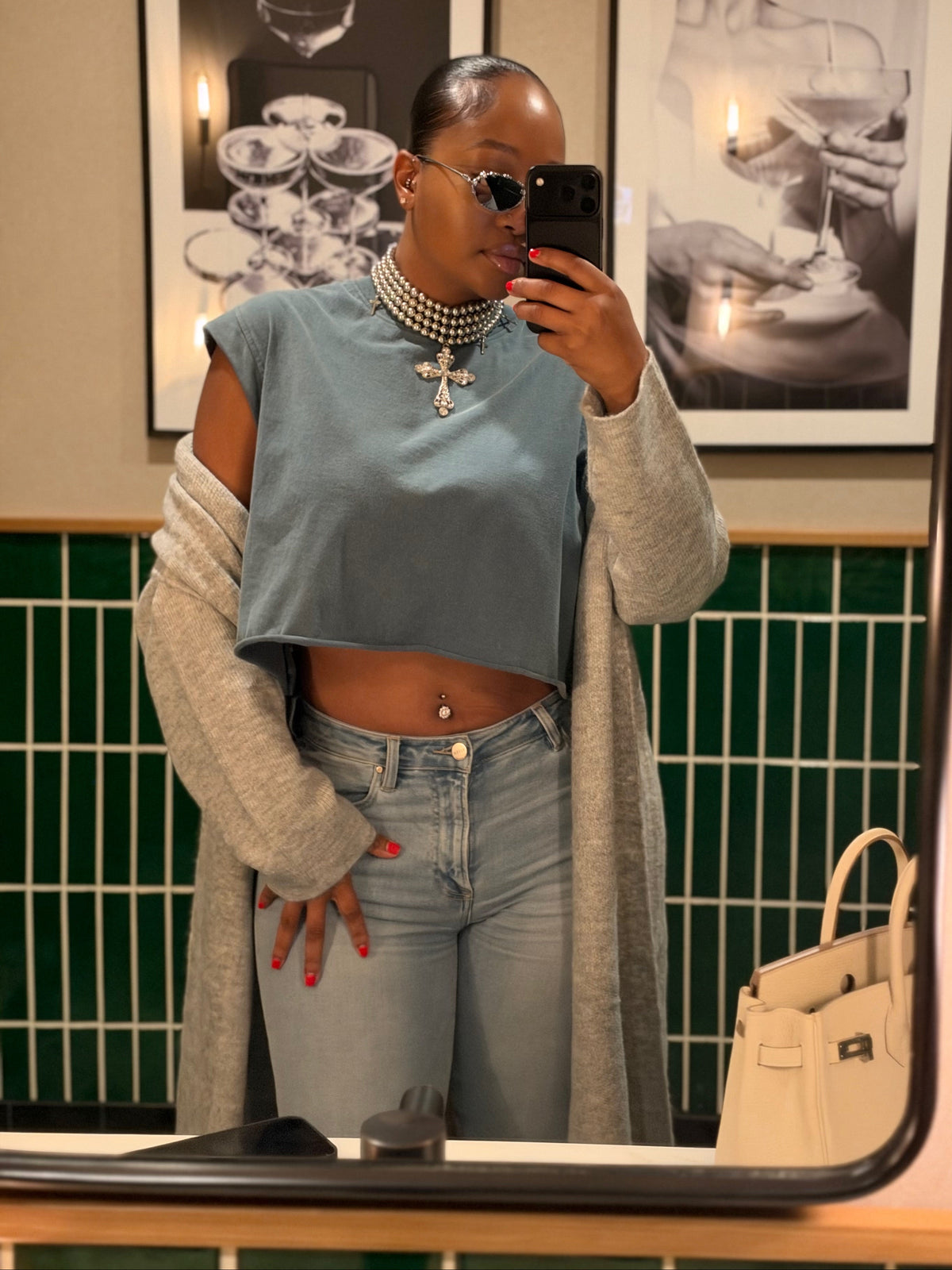 A woman takes a mirror selfie showing off a light blue cropped top with a gray cardigan, light-wash jeans, and a pearl necklace. The background features a green tiled wall and framed black-and-white art.