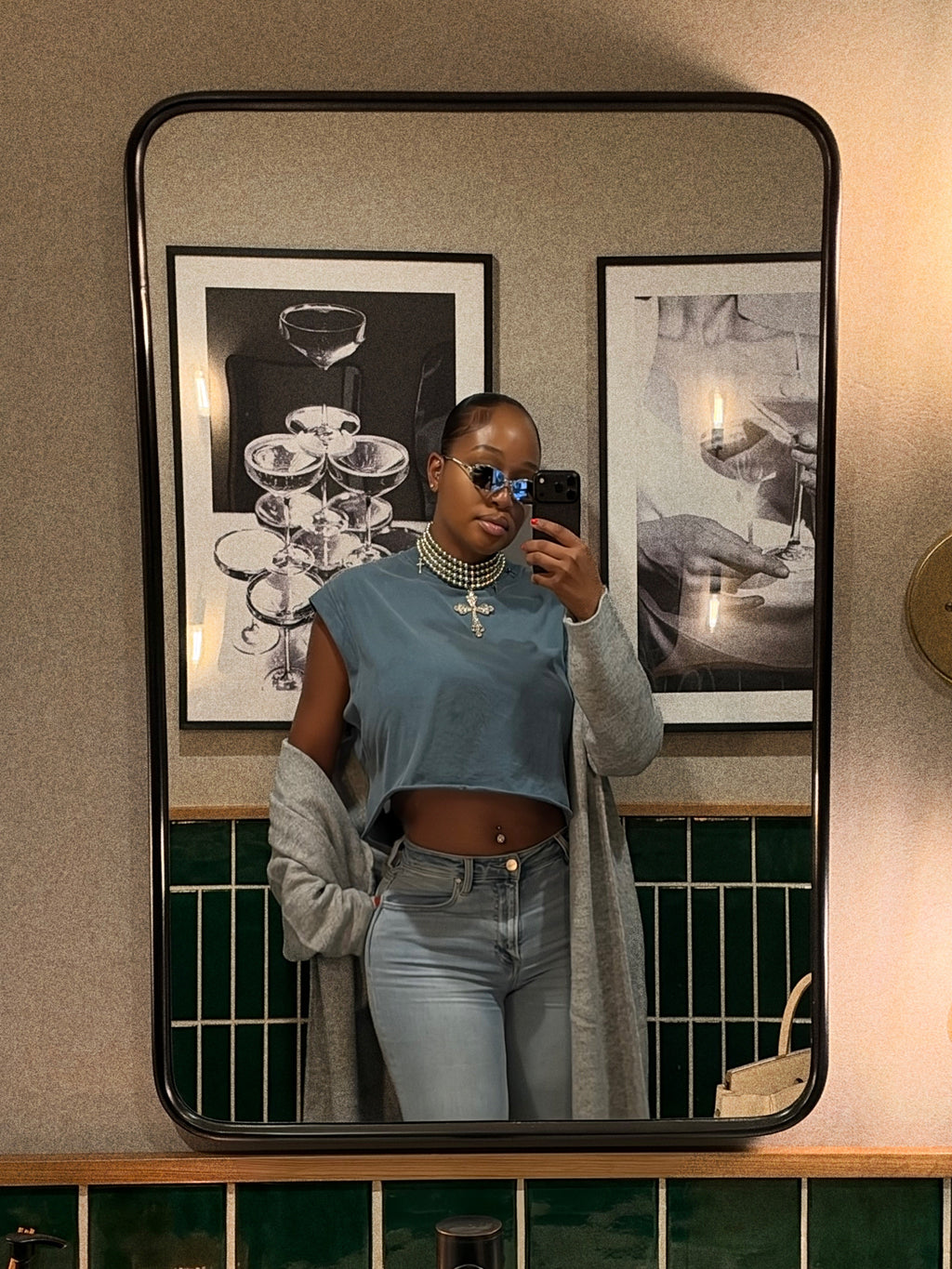 A woman taking a mirror selfie wearing a light blue cropped top, a gray cardigan, and light-wash jeans. She is wearing blue-tinted sunglasses and a cross necklace.