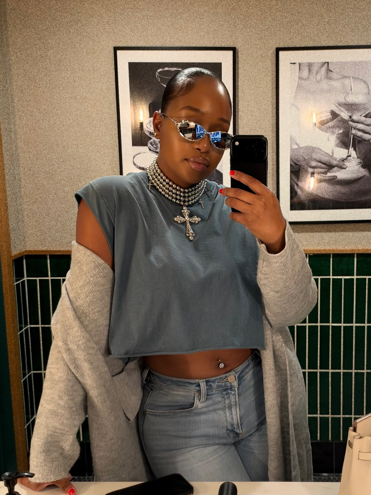 A woman taking a mirror selfie wearing a slate blue cropped top with cold-shoulder detailing, a light gray cardigan, light-wash jeans, layered pearl necklaces, and silver rhinestone sunglasses.