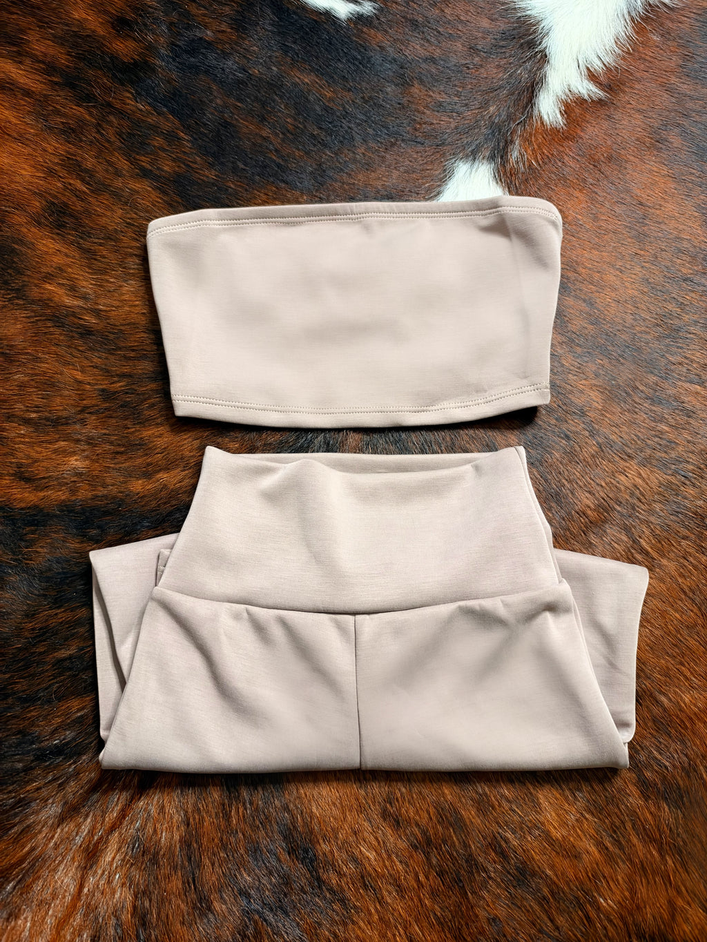 A two-piece matching set of light beige athletic wear, including a strapless bandeau top and high-waisted leggings, laid flat on a brown and black cowhide rug.