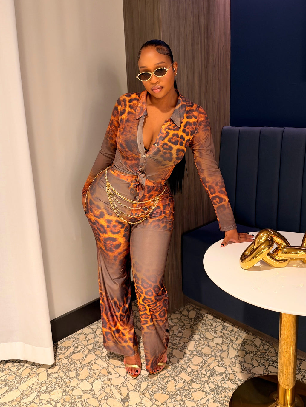 A woman posing in a stylish indoor setting wearing a sheer, long-sleeve jumpsuit with an orange and brown leopard print pattern over a gray base. She is standing next to a white side table with a gold chain sculpture and a blue tufted bench.