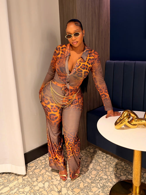 A woman posing in a stylish indoor setting wearing a sheer, long-sleeve jumpsuit with an orange and brown leopard print pattern over a gray base. She is standing next to a white side table with a gold chain sculpture and a blue tufted bench.