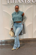 A woman posing in a light blue sleeveless crop top, light-wash distressed flare jeans, a gray cardigan, and patterned pointed-toe flats, holding a cream-colored handbag against a white textured wall.